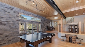 A spacious room with a ping pong table, stone wall, wooden ceiling, and large windows overlooking a natural landscape. - Golden Oak Estate by Destination Big Bear : Luxury Log Home in Castle Glen! Walk to the Lake! Beautiful Mountain Views! (Big Bear Lake)