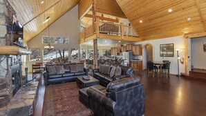 A spacious living area with a high ceiling, wooden beams, a fireplace, and a view of the outdoors. - Golden Oak Estate by Destination Big Bear : Luxury Log Home in Castle Glen! Walk to the Lake! Beautiful Mountain Views! (Big Bear Lake)