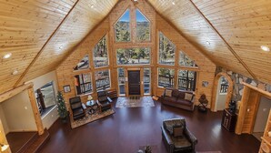 A spacious loft-style room with a high ceiling, large windows, and a cozy seating area with leather sofas and a coffee table. - Golden Oak Estate by Destination Big Bear : Luxury Log Home in Castle Glen! Walk to the Lake! Beautiful Mountain Views! (Big Bear Lake)