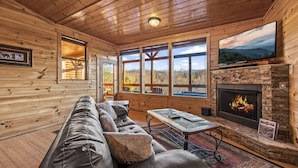 A log cabin interior with a stone fireplace, a large TV, and a sofa. - Love Me Tender: Amazing Views, Romantic, Fire Pit (Gatlinburg)