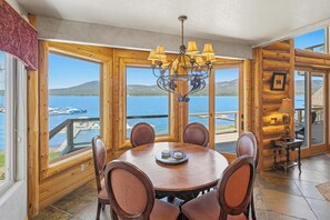 Een houten interieur met een ronde tafel en stoelen, grote ramen met uitzicht op een meer en boten, en een kroonluchter.