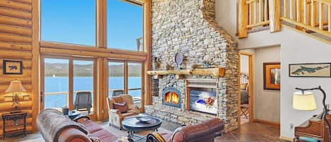 A log cabin interior with a stone fireplace, leather sofas, and a view of a lake.