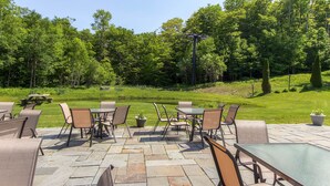 An outdoor patio area with stone flooring, multiple tables and chairs, and a view of a lush green landscape with trees.