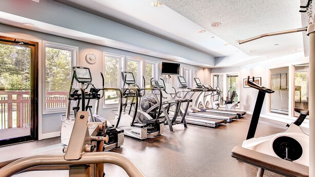 A gym with various cardio machines, large windows, and a view of the outdoors.