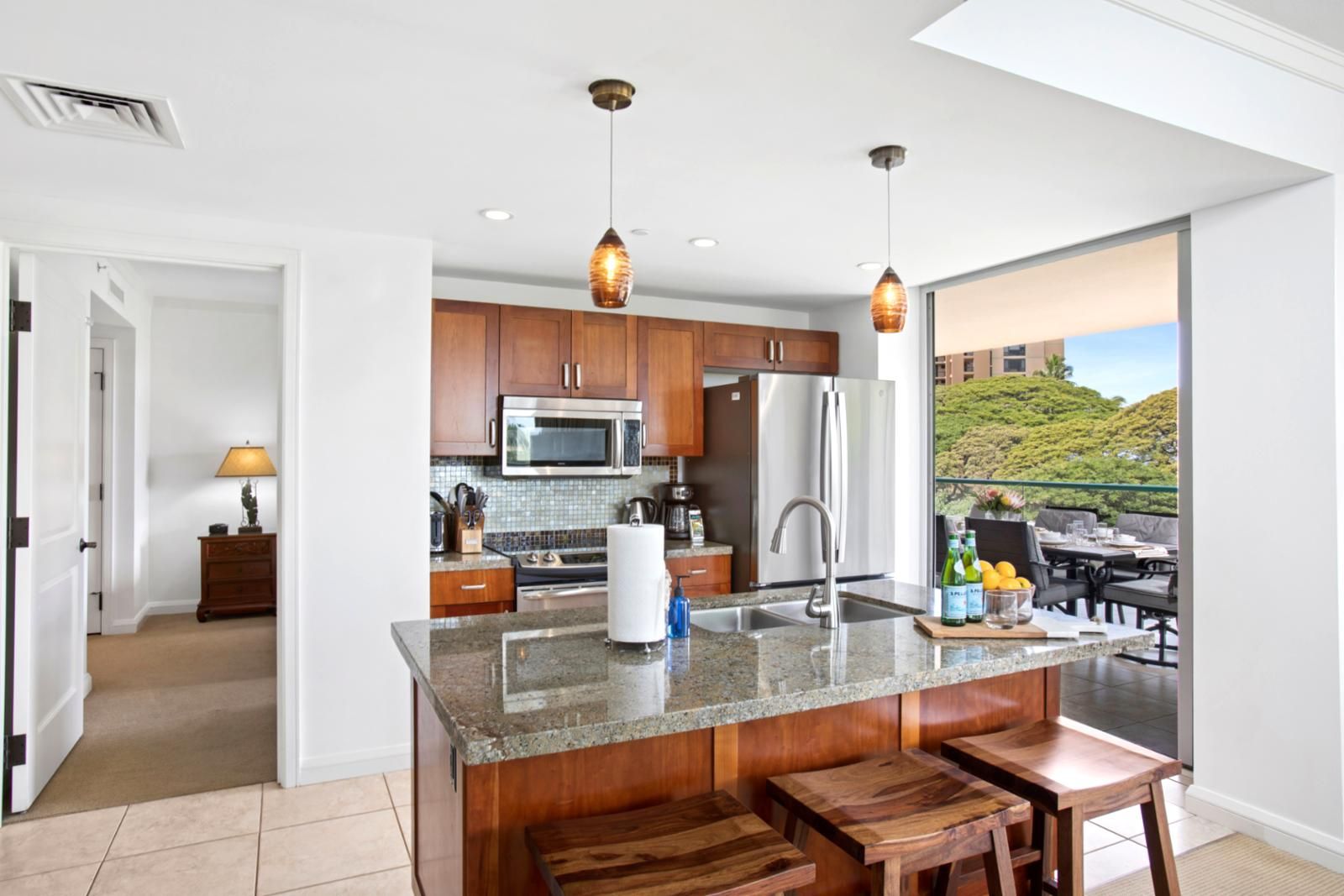 Una cocina moderna con encimeras de granito, electrodomésticos de acero inoxidable y armarios de madera. Cuenta con una isla grande con taburetes de bar y vistas a un paisaje exuberante a través de una puerta corrediza de vidrio.