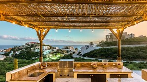 Cozinha externa com churrasqueira, pia e espaço de bar, com vista para a paisagem costeira com edifícios e vegetação.