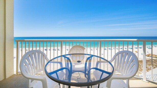 Un balcon avec une table en verre et deux chaises, donnant sur une plage bordée d’un ciel bleu azur et d’un rivage sablonneux.