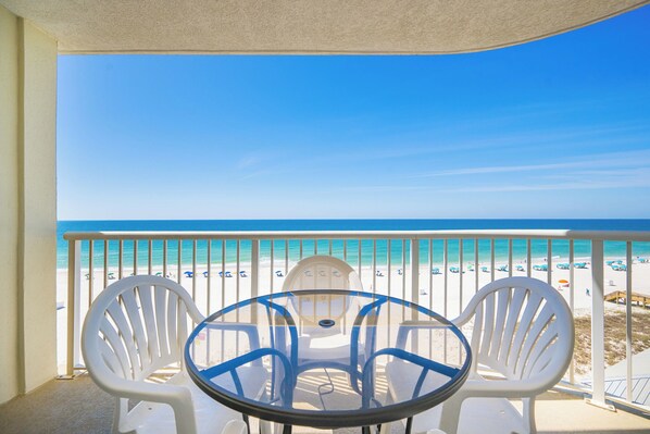 Varanda com mesa de vidro e duas cadeiras, vista para uma praia com céu azul límpido e faixa de areia.