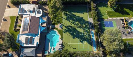 An aerial view of a residential property with a swimming pool, tennis courts, and a solar panel-equipped house.