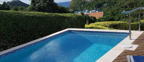 A rectangular swimming pool with a wooden deck and metal railings, surrounded by neatly trimmed hedges and a mountainous backdrop.