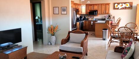A modern living room with a flat-screen TV, wicker furniture, and a dining area with wooden chairs and a kitchen in the background.
