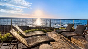 A balcony with wicker lounge chairs overlooking a body of water, a railing, and a clear sky.