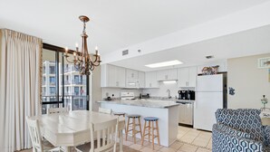 A modern kitchen with white cabinets, a central island, and bar stools. There is a dining table with chairs and a chandelier. A patterned sofa is visible in the background. - Phoenix VII 301 | Beachfront | Updated | Pools | My Beach Getaways (Orange Beach)