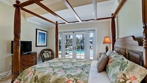 A four-poster bed with a floral bedspread, a television on a wooden dresser, and a view of a pool through glass doors. - 510 Bayview -  Anna Maria Island - Private home 5 Bedroom / 5 Bath with private pool  , maximum occupancy of 6 people. (Holmes Beach)