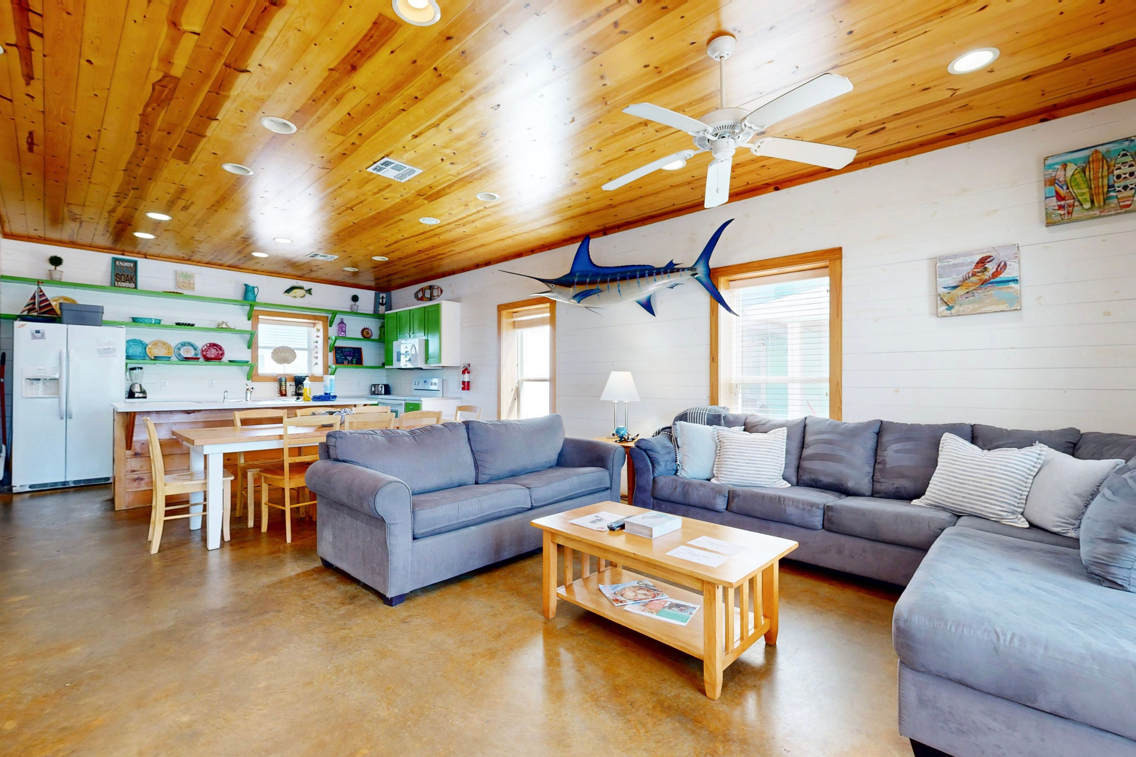 A spacious living area with a wooden ceiling, a dining table, two sofas, and a kitchen in the background.