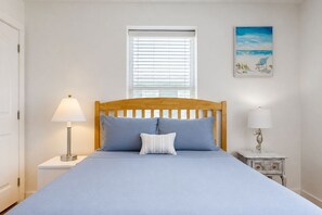 A bedroom with a bed, a nightstand, a lamp, a window with blinds, and a painting on the wall.
