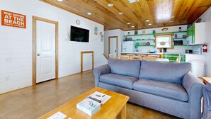 Ein Zimmer mit grauem Sofa, Holzboden, weißen Wänden, einem Fernseher, einer Uhr und einer Küchenzeile mit grünen Schränken.