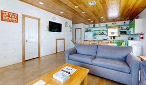 A room with a grey sofa, wooden ceiling, white walls, a television, a clock, and a kitchen area with green cabinets. - Pet Friendly, Close to beach, Shared Pool, Golf Cart Access (Port Aransas)