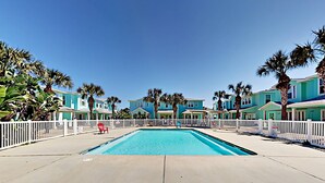 Ein Poolbereich mit blauem Himmel, umgeben von zweistöckigen Gebäuden mit weißen Zäunen, Palmen und einem roten Stuhl.