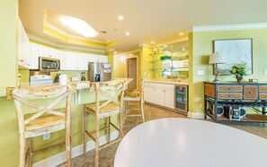Una cocina con armarios blancos, microondas, refrigerador y cafetera. Hay un comedor con mesa y sillas blancas.