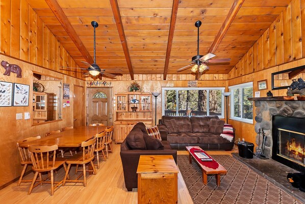 A cozy cabin interior with wooden walls, a dining area, a living room with a fireplace, and a ceiling fan.