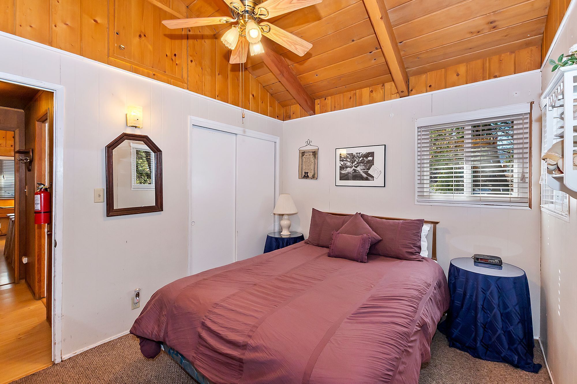 Un dormitorio con techo de madera, una cama con colcha rosa, una mesita de noche, una lámpara, un cuadro enmarcado y una ventana con persianas.