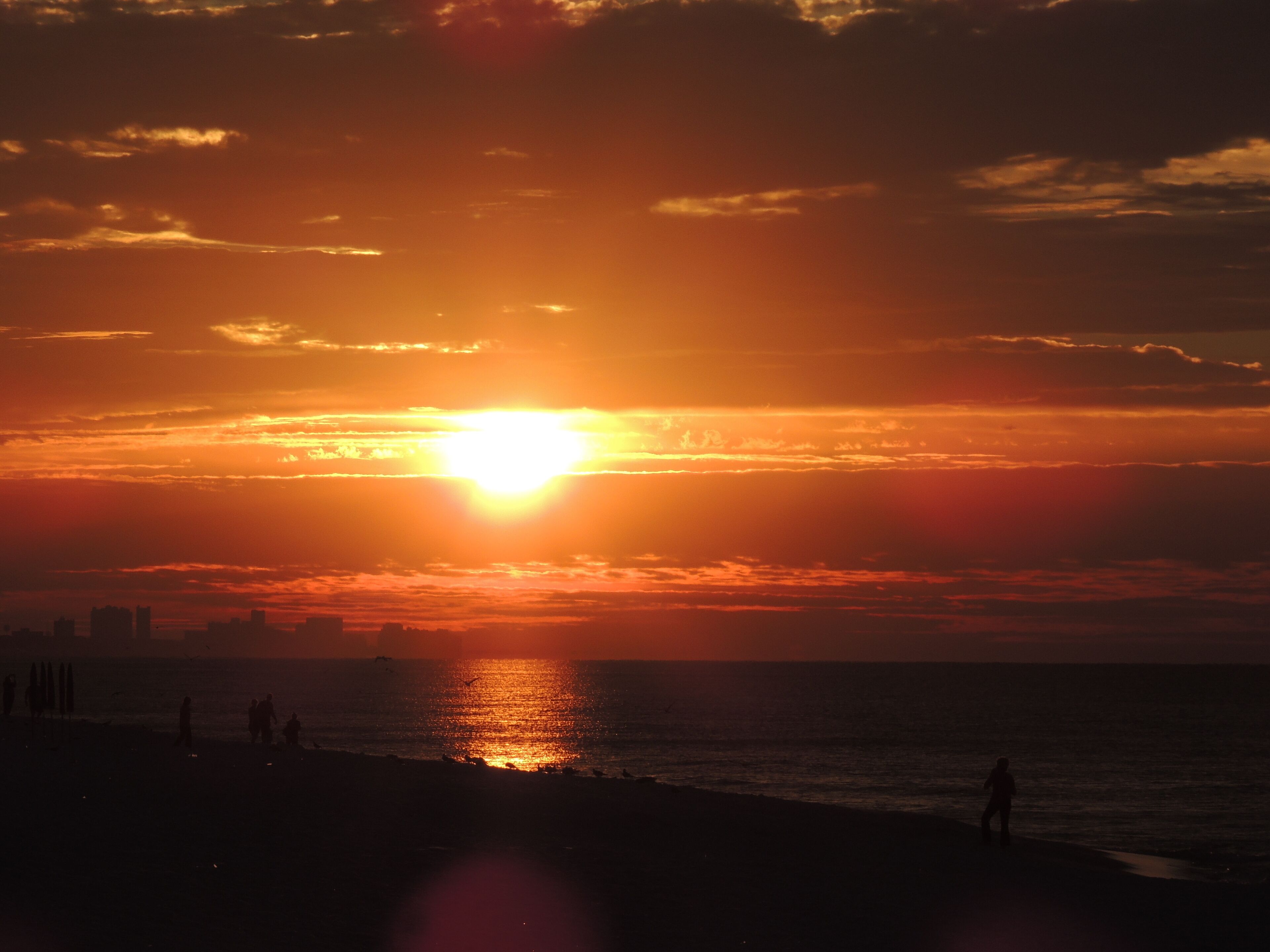 A sunset over a beach with silhouettes of people and buildings in the distance.