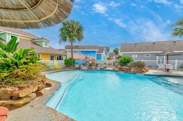 A swimming pool with a rock feature, surrounded by colorful buildings and palm trees.