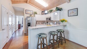 Une cuisine avec des meubles blancs, un îlot central doté de tabourets, des poutres en bois au plafond et des luminaires suspendus.