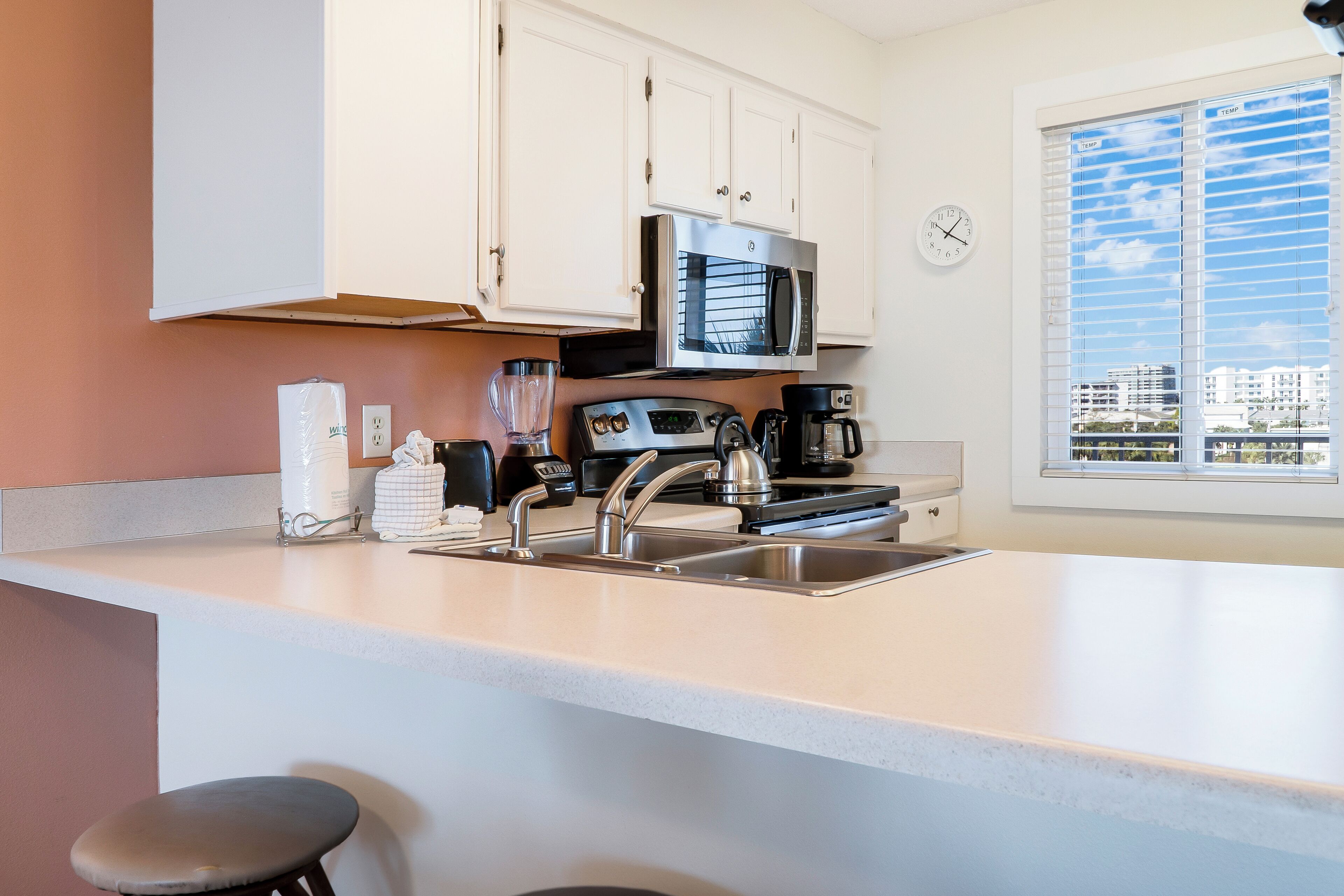 Een moderne keuken met witte kasten, een roestvrijstalen spoelbak, een magnetron, een koffiezetapparaat en een raam met uitzicht op gebouwen.