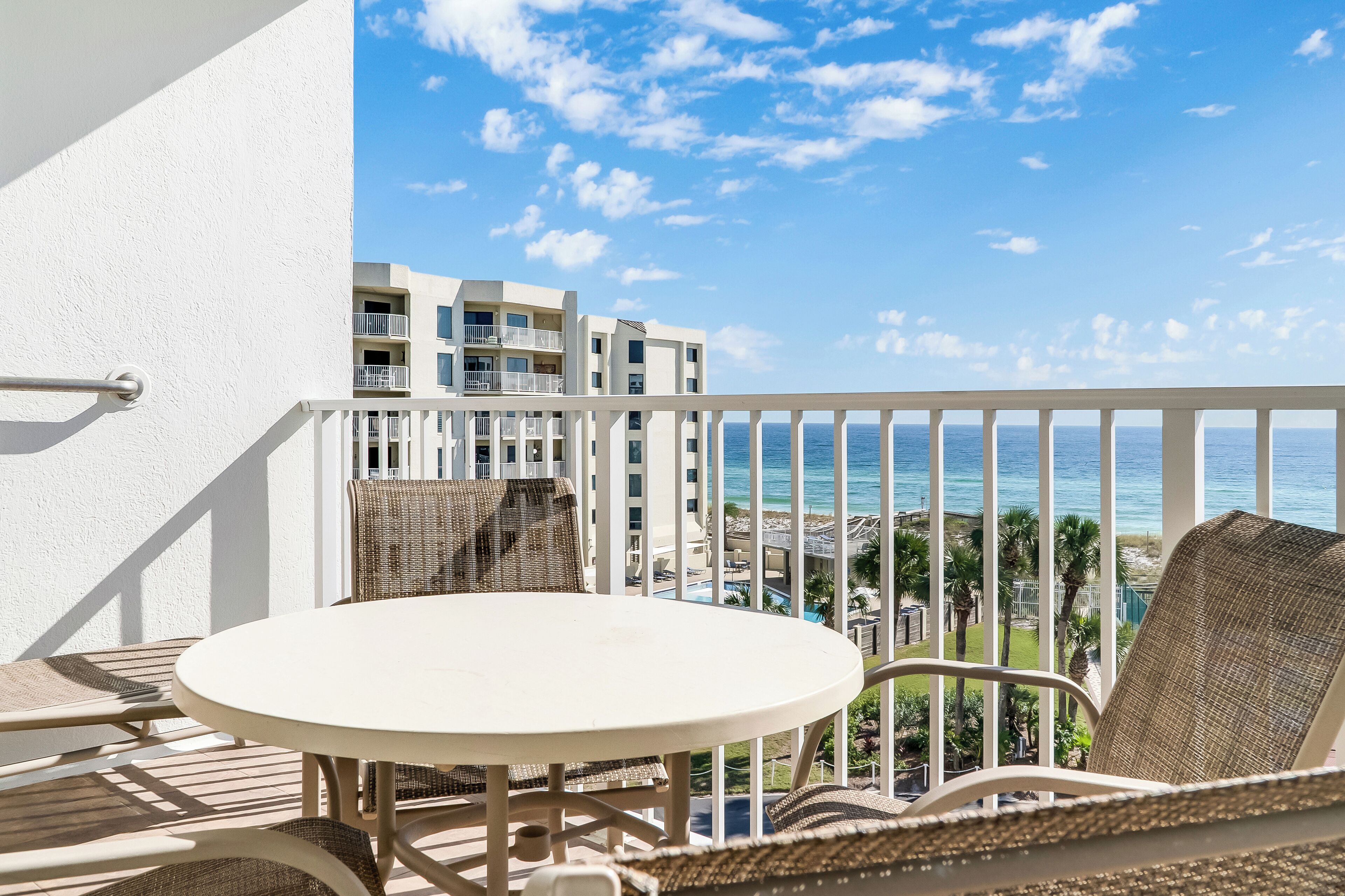 A balcony with a round table and wicker chairs, overlooking a resort area with buildings and the ocean.