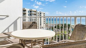 A balcony with a round table and wicker chairs, overlooking a resort area with buildings and the ocean.
