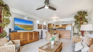 A spacious living area with a large flat-screen TV, wicker furniture, and a kitchen in the background.
