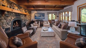 Un salon chaleureux avec une cheminée en pierre, des sièges confortables, un téléviseur à écran plat et de grandes fenêtres donnant sur l’extérieur.