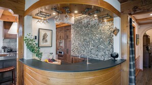 A kitchen with a stone wall, wooden cabinets, a black countertop, and a hanging wine rack.