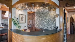 Une cuisine avec un mur en pierre, des meubles en bois, un plan de travail noir et un porte-bouteilles à vin suspendu.