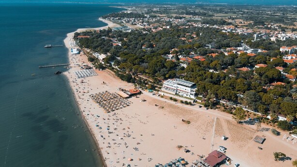Vue aérienne d’une ville côtière avec une plage de sable, une jetée et un grand bâtiment doté d’un toit rouge.
