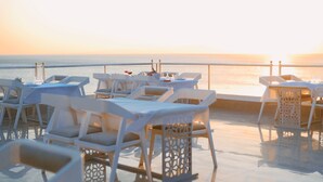 Une terrasse avec des chaises et des tables en plastique blanc, donnant sur une mer calme au coucher du soleil.