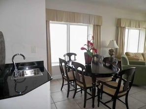 A dining area with a black countertop, a sink, and a table with chairs. There is a window with curtains and a green sofa in the background.