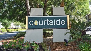 Un letrero con la palabra 'Courtside', rodeado de follaje y una carretera.