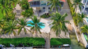 An aerial view of a resort with a swimming pool, palm trees, and a dock.