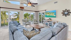 A spacious living room with a large gray sectional sofa, a wooden coffee table, and a ceiling fan. There is a large window offering a view of palm trees and a building outside.