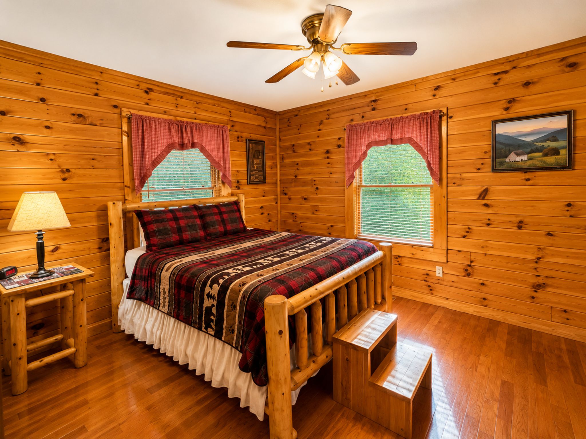 Un dormitorio de cabaña de madera con una cama, una mesita de noche, una lámpara, un ventilador de techo y dos ventanas con cortinas.