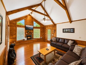 A spacious living room with wooden floors, a fireplace, a ceiling fan, and a large window offering a view of the outdoors.