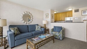 Una sala de estar con un sofá azul, una mesa de centro de vidrio y un sillón estampado. Se ve una cocina al fondo con armarios de madera y un microondas.