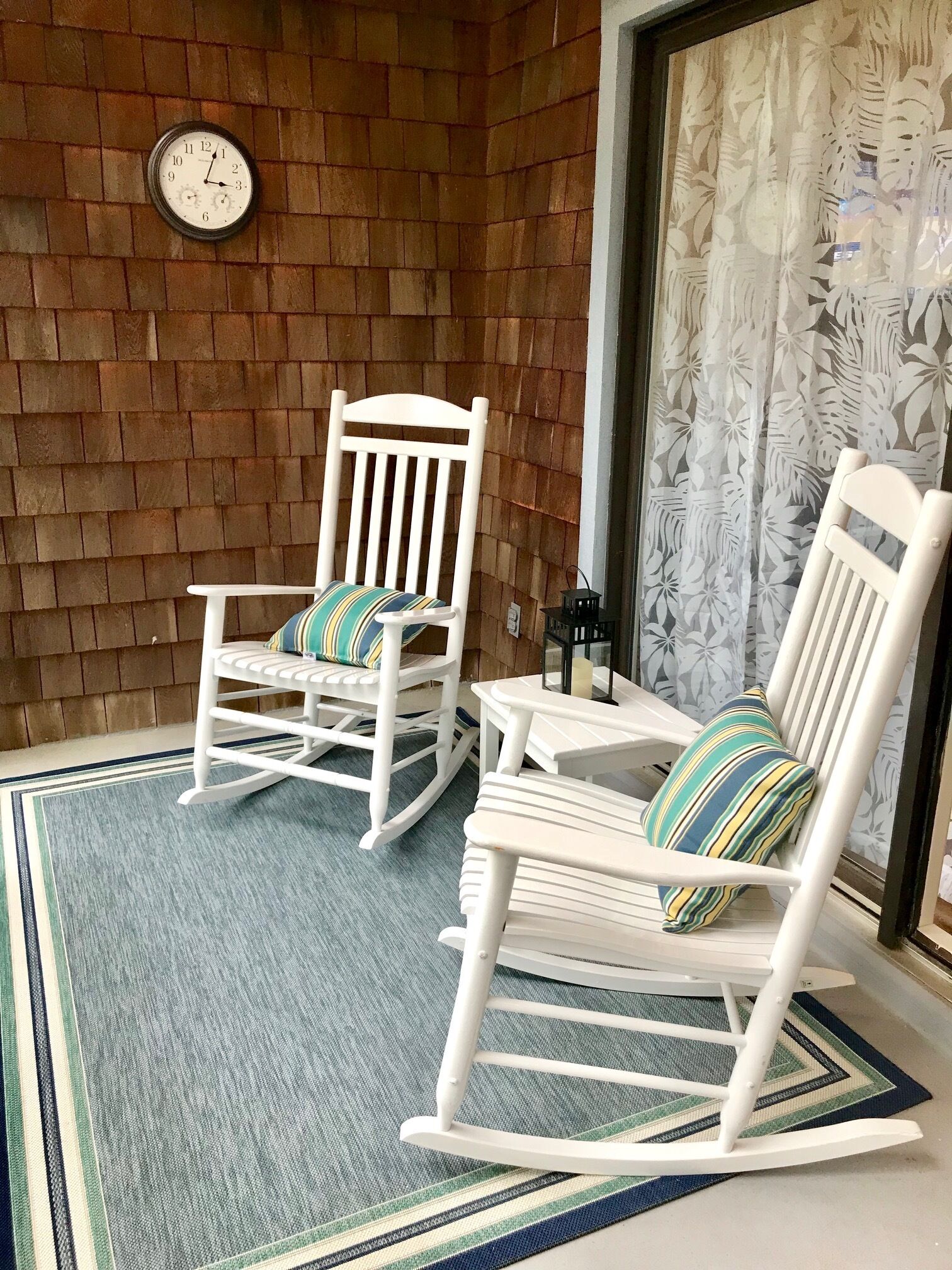 Un patio con dos mecedoras y una pequeña mesa, un reloj en la pared y una cortina estampada.