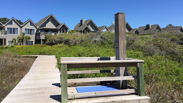 Un paseo marítimo de madera que lleva a una casa de playa con un felpudo azul en la terraza.