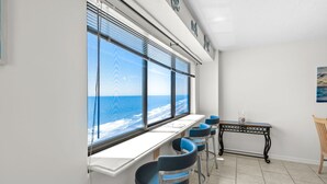 A room with a large window overlooking the sea, a white counter, blue chairs, a small table, and a wooden chair.