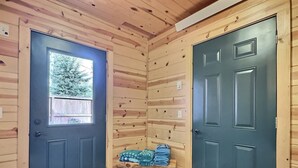 Ein Interieur einer Holzhütte mit zwei dunklen Türen, einer kleinen Bank mit zusammengeklappten Handtüchern und einem Fenster mit Blick auf Bäume.