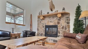 Ein gemütliches Wohnzimmer mit Kamin aus Stein, einem Ledersofa, einem Holztisch und einem großen Fenster.